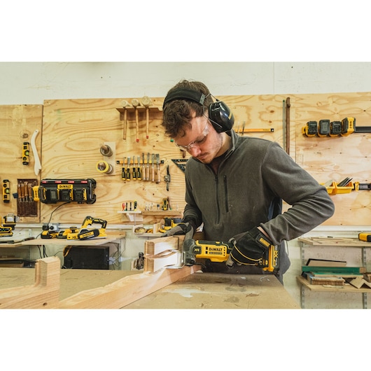 Person using a DEWALT DCS359N-XJ power tool on wood in a workshop with various tools, wearing safety gear.