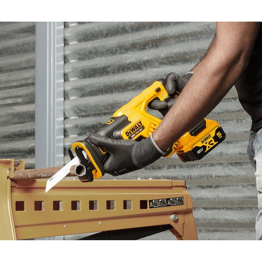 A person wearing gloves is using a yellow and black DEWALT cordless reciprocating saw (model DCS367P2) to cut through a metal pipe placed on a workbench.
