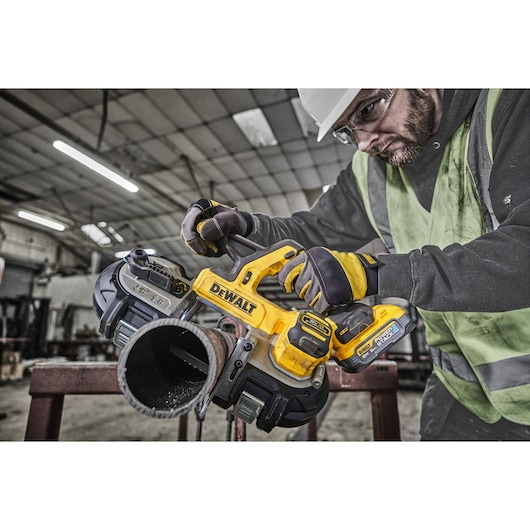 A DEWALT cordless handheld band saw (SKU: DCS378N) cutting through a metal pipe in an industrial setting. The tool is yellow and black and held by a person wearing gloves and a safety vest. The background shows a large workspace with industrial equipment.