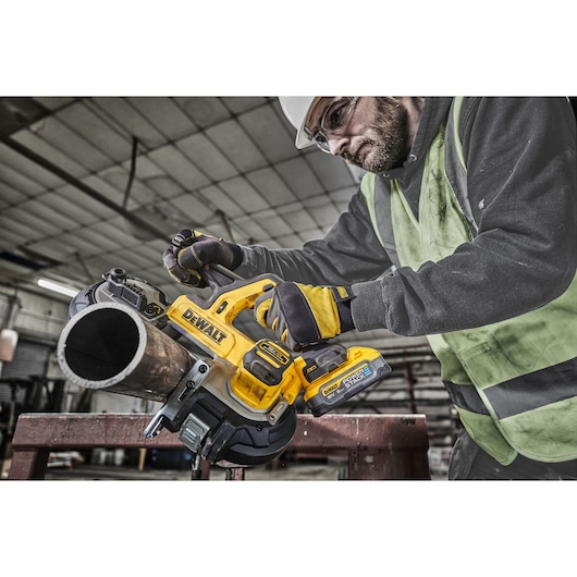 A person wearing work gloves and a safety vest is using a DEWALT DCS378N cordless band saw to cut through a metal pipe inside an industrial workshop.