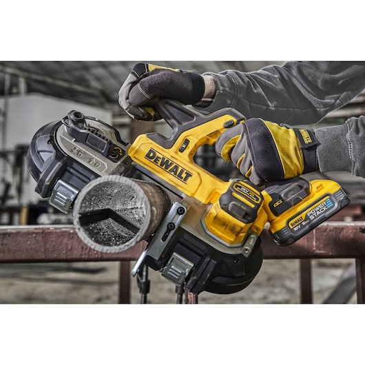 A person wearing protective gloves is using a DEWALT cordless band saw to cut through a metal pipe in an industrial workshop setting.