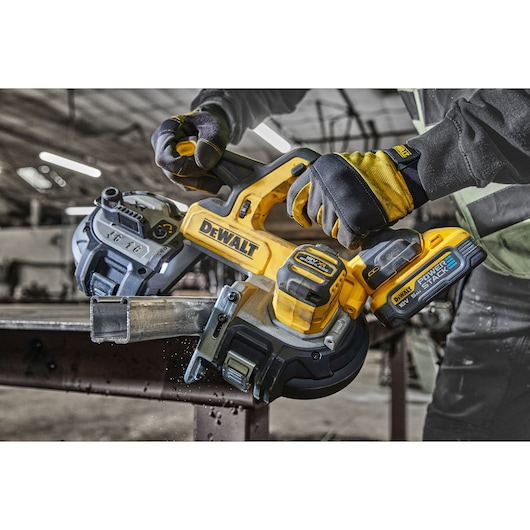 A person wearing protective gloves uses a DEWALT DCS378N cordless band saw with a POWERSTACK battery to cut a metal pipe in an industrial workshop.
