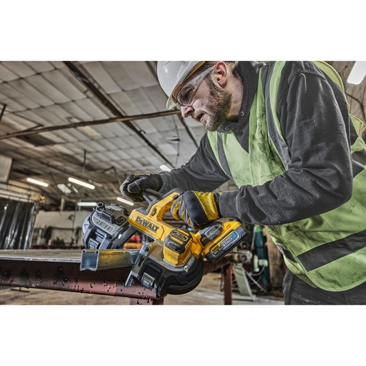 A person wearing a safety vest and gloves is operating a DEWALT cordless band saw (model DCS378N) to cut a metal beam in an industrial workshop.