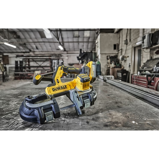 A yellow and black DEWALT cordless band saw placed on a metal workbench in an industrial workshop setting. The tool appears ready for use, surrounded by various equipment and materials in the background.