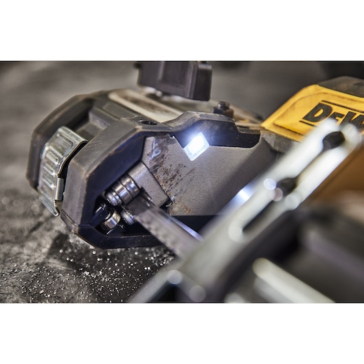Close-up view of a DEWALT portable band saw cutting through metal, with visible LED light illuminating the cutting area. The saw blade and metal surface are shown with metal shavings.