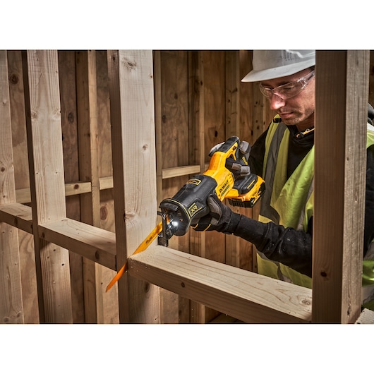 A person wearing safety gear uses a DEWALT cordless reciprocating saw to cut through a wooden beam in a construction setting.