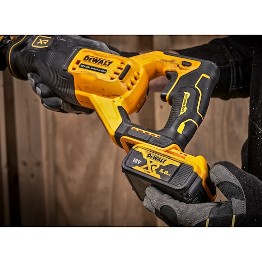 Close-up of a person holding a DEWALT 18V XR brushless cordless reciprocating saw with an attached 5.0Ah lithium battery, wearing work gloves, in front of a wooden background.