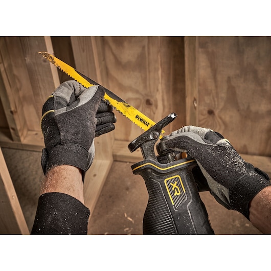 Close-up of gloved hands inserting a yellow DEWALT saw blade into a black reciprocating saw, with wooden framing in the background.