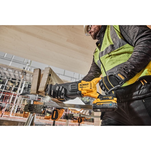 A construction worker using a DEWALT DCS386H2T cordless reciprocating saw to cut a piece of wood at a building site, wearing safety gear including gloves, vest, and helmet.