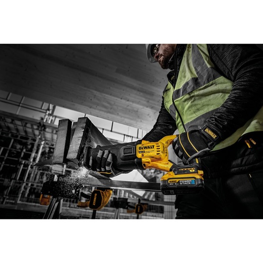 A person wearing safety gear uses a DEWALT cordless reciprocating saw, model DCS386H2T, with POWERSTACK technology to cut through a metal beam in a construction setting.