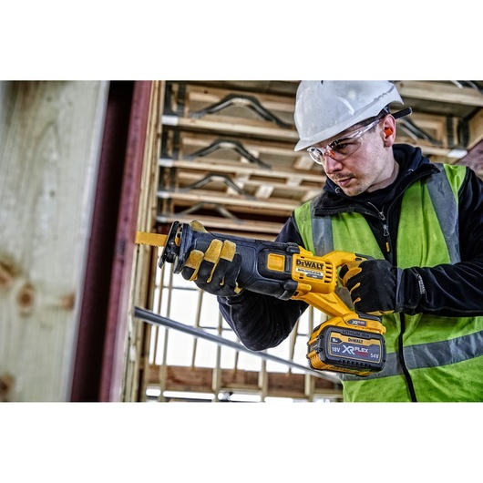 Person using a DEWALT DCS389N cordless reciprocating saw at a construction site with safety gear.