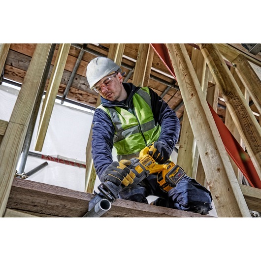 Person using a DEWALT cordless saw to cut pipe at a construction site, wearing safety gear and high-visibility vest.