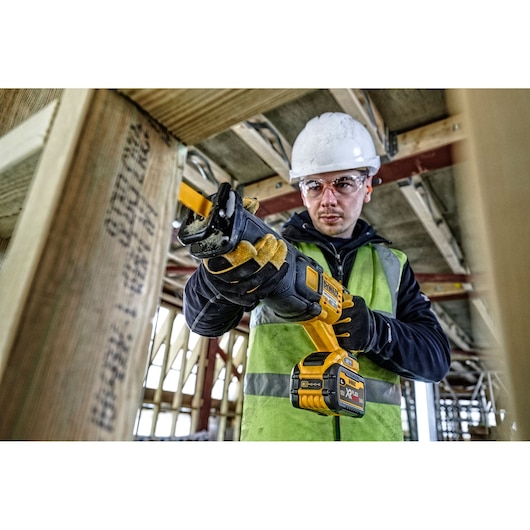 A construction worker using a DEWALT cordless reciprocating saw (model DCS389T2) to cut through a wooden beam at a building site. The worker is wearing protective gloves, a safety vest, and a hard hat.