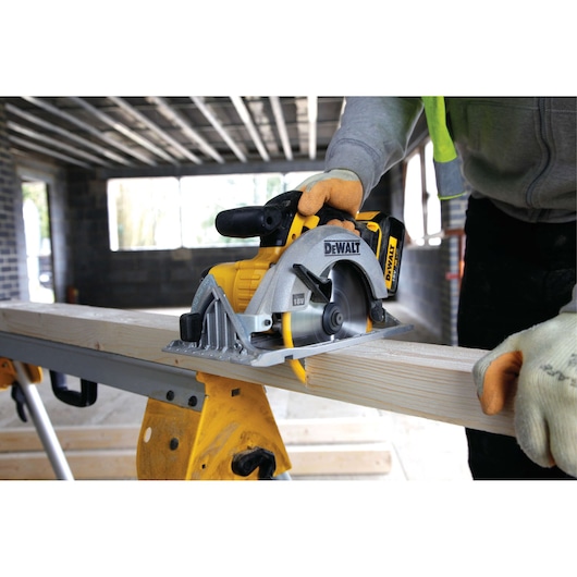 A person wearing safety gloves uses a DEWALT 18V cordless circular saw to cut a wooden plank on a workbench in a construction setting.