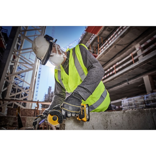 A construction worker wearing a safety vest, helmet, gloves, and face shield is using a DEWALT cordless angle grinder on a concrete surface at an outdoor construction site.