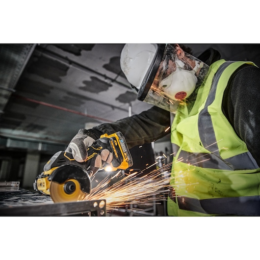 A person wearing a safety helmet, face shield, mask, gloves, and a high-visibility vest is using a DEWALT cordless angle grinder (model DCS438N) to cut metal, causing sparks to fly.