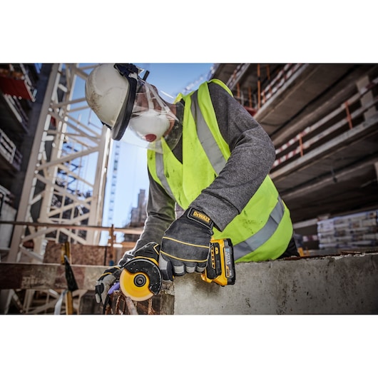 A construction worker wearing a high-visibility vest, helmet, protective face shield, and gloves is using a DEWALT DCS438N power tool to cut through concrete at a construction site.