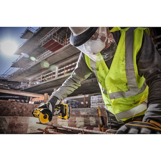 A construction worker wearing a high-visibility vest, safety gloves, face shield, and mask is using a DEWALT DCS438N cordless tool to cut metal rods on a building site.