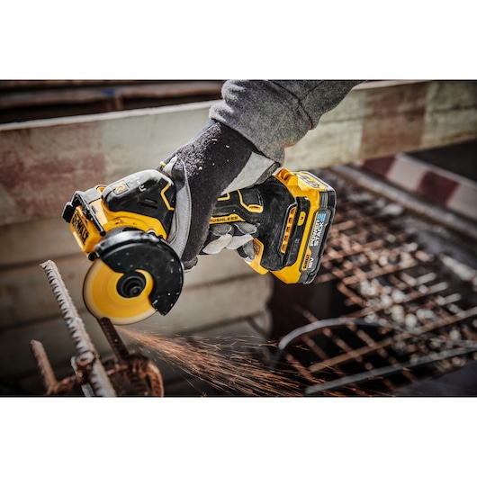 A person wearing gloves is using a yellow DEWALT DCS438N cordless brushless angle grinder to cut through a metal rod, while sparks fly from the cutting area.