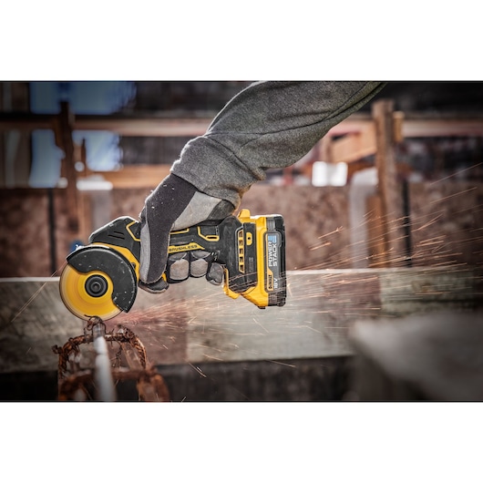 A person wearing work gloves uses a DEWALT cordless brushless angle grinder with a yellow and black design to cut through a metal rod, causing sparks to fly.
