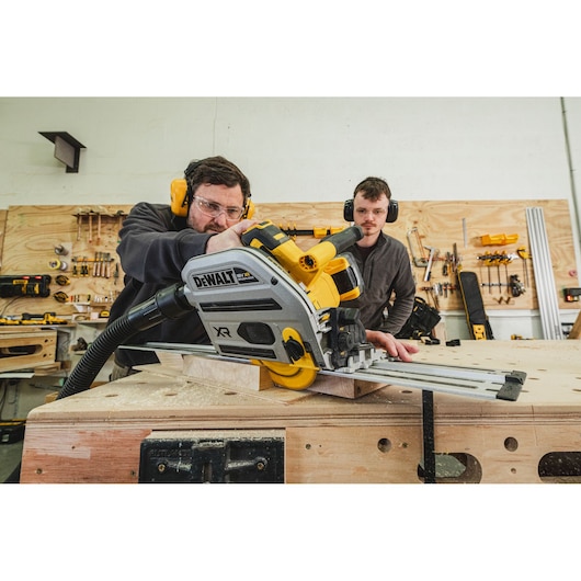 Two people using a DEWALT cordless track saw to cut wood in a workshop with tools on the wall.