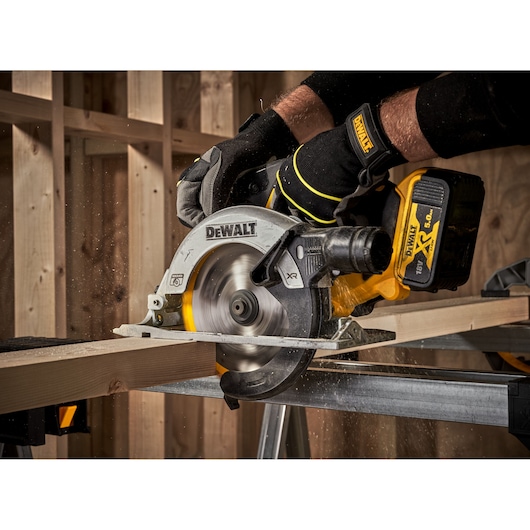 A DEWALT cordless circular saw being used to cut a piece of wood, operated by a person wearing protective gloves. The saw is in action in a workshop setting.