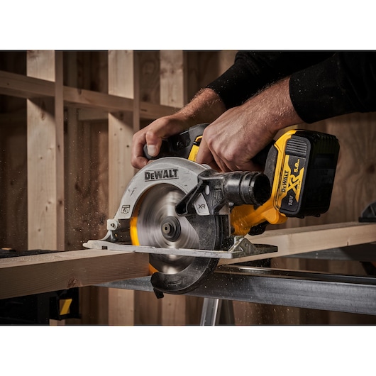 A close-up of a person using a DEWALT cordless circular saw to cut a wooden plank inside a construction site.
