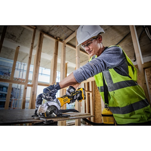 A construction worker wearing a safety vest, gloves, and a helmet uses a DEWALT cordless circular saw (model DCS571E1T) to cut a piece of wood at a building site with exposed framing.