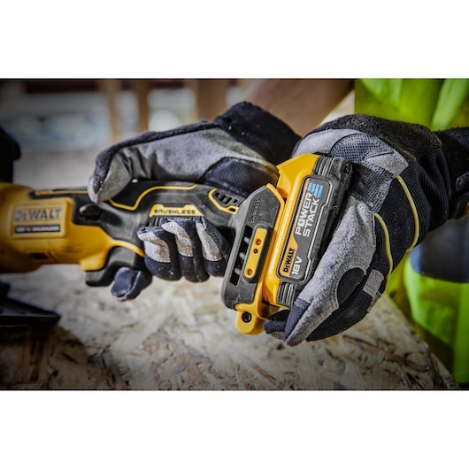 Close-up of a person wearing work gloves attaching an 18V DEWALT POWERSTACK battery to a DEWALT brushless power tool on a wooden surface.