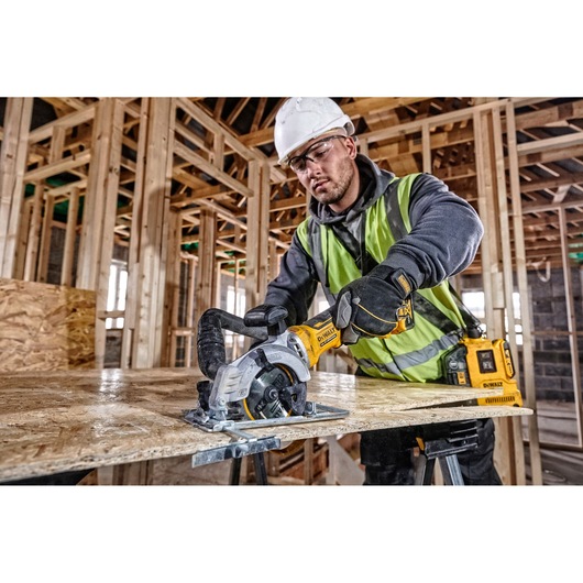 Person uses DEWALT DCS571N saw to cut wood sheet at construction site, wearing safety gear and high-vis vest.