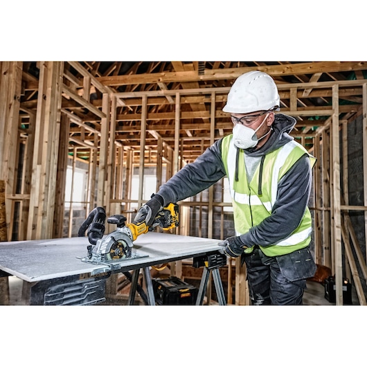 Worker uses a DEWALT cordless circular saw to cut a board at a construction site with wooden framing.