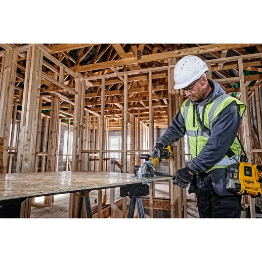 Worker uses DEWALT DCS571N saw to cut wood panel at construction site, wearing safety gear and high-visibility vest.