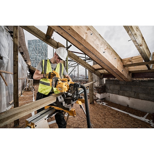 A construction worker wearing a high-visibility vest and a hard hat operates a DEWALT cordless mitre saw (SKU: DCS777N) to cut a wooden beam on a construction site. The surroundings include partially built structures with exposed beams and urban buildings in the background.