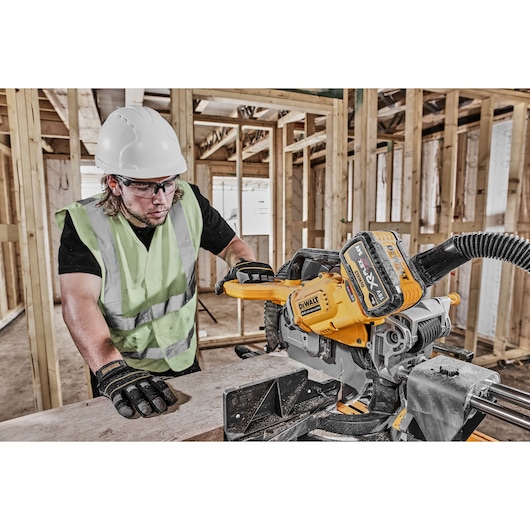 A worker wearing a safety helmet, high-visibility vest, and gloves operating a DEWALT DCS781N power tool, likely a cordless miter saw, on a construction site with wooden framing in the background.