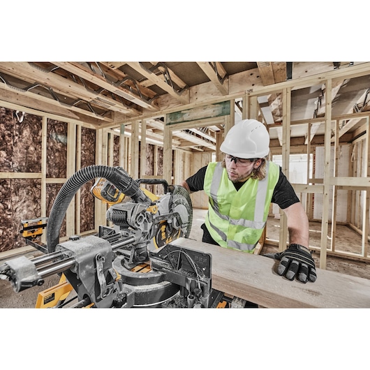 A person wearing a hard hat, high-visibility vest, and gloves operates a DEWALT cordless mitre saw to cut a piece of wood in a building under construction. The construction site features exposed wooden framework and insulation.