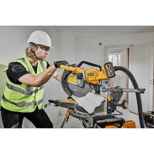 A person wearing a safety vest, hard hat, and gloves is operating a DEWALT DCS781N cordless mitre saw in a construction setting. The saw is cutting a piece of white trim, and a dust extraction hose is attached.