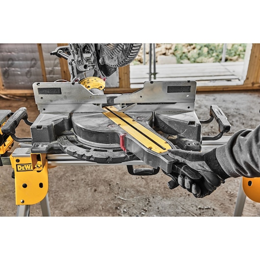 A DEWALT miter saw (model DCS781N) is shown in use at a workshop, with a gloved hand adjusting the saw's angles. The tool is mounted on a yellow stand and positioned for precise wood cutting.
