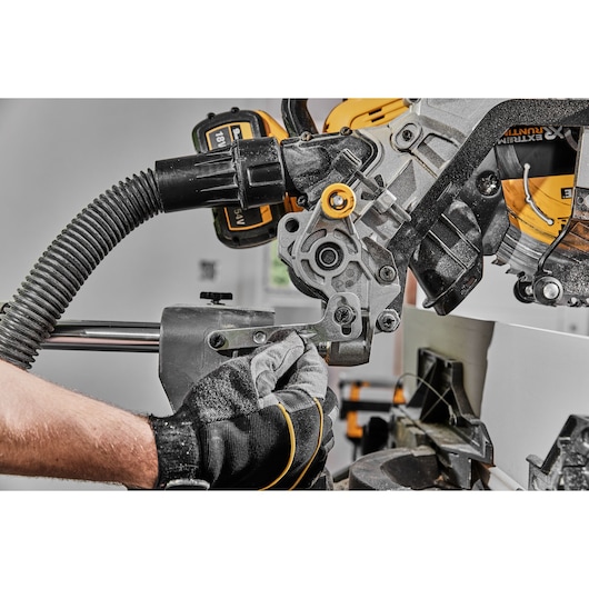 Close-up of a person wearing gloves adjusting a DEWALT DCS781N power tool, possibly a compound miter saw, in an indoor workshop setting.