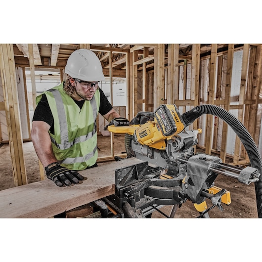 A construction worker wearing a high-visibility vest, gloves, and a hard hat is using a DEWALT DCS781X2 cordless miter saw to cut a wooden board at a construction site. The background shows the wooden frame of an unfinished building.