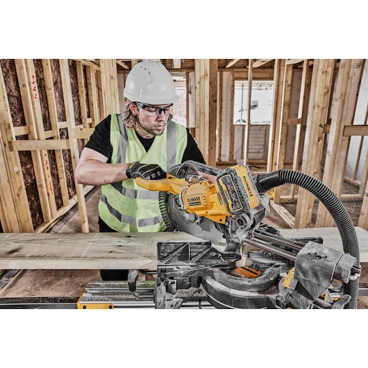 A worker wearing a safety helmet and high-visibility vest uses a DEWALT DCS781X2 cordless miter saw to cut a piece of wood at a construction site. The tool is connected to a dust extraction hose.