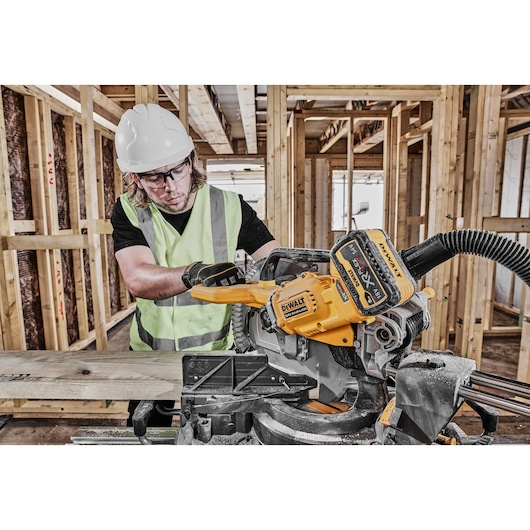 A person wearing a safety vest, gloves, and a hard hat using a DEWALT DCS781X2 cordless miter saw to cut a wooden plank at a construction site with unfinished wooden framing.