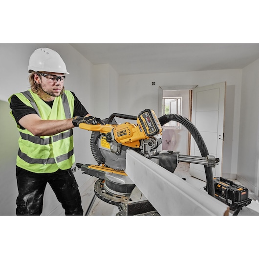 A construction worker wearing a white hard hat and reflective vest operates a DEWALT DCS781X2 cordless miter saw to cut a piece of wood indoors. The saw, with a connected dust extraction hose, is positioned on a workbench in a room under renovation.