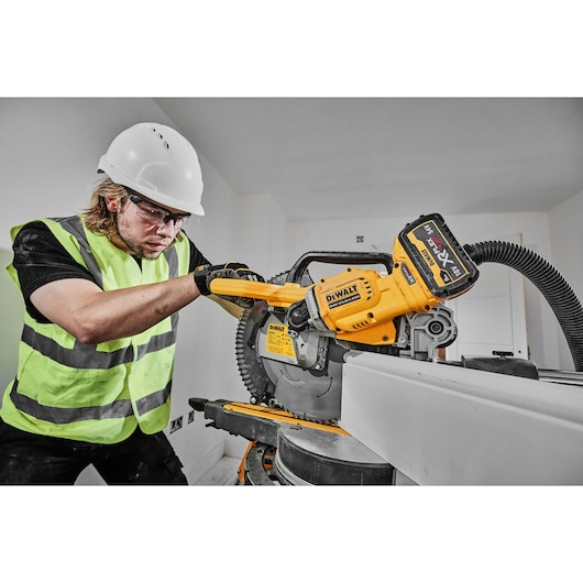 A person wearing a safety helmet and high-visibility vest is operating a DEWALT DCS781X2 miter saw in an indoor construction setting.