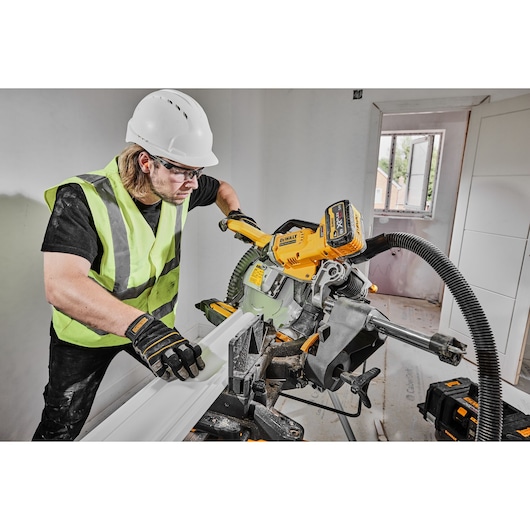 A person wearing a safety vest, gloves, and hard hat is using a DEWALT DCS781X2 cordless miter saw to cut a piece of trim inside a room under construction. The saw is connected to a dust extraction system, and there are DEWALT tool batteries visible nearby.
