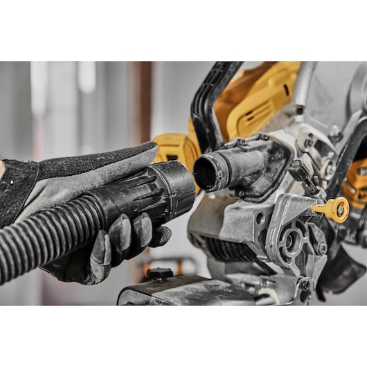 A close-up image shows a gloved hand connecting a vacuum hose to a DEWALT power tool, specifically a miter saw, to help with dust extraction.