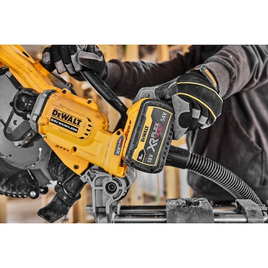 Close-up of a person wearing gloves using a DEWALT brushless saw, specifically inserting or removing a DEWALT XR FLEXVOLT 18V/54V battery. The yellow and black color scheme, branding, and construction environment are visible.