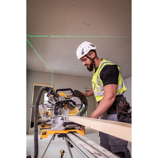 A construction worker wearing a safety helmet and high-visibility vest operates a DEWALT DCS782N saw to cut a wooden plank indoors.