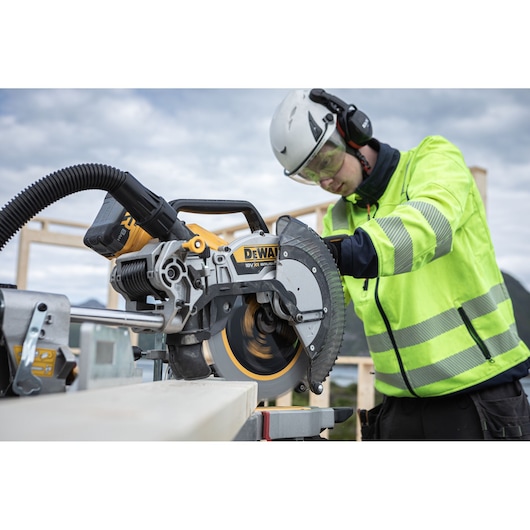 A person wearing safety gear uses a DEWALT DCS782N miter saw to cut wood at a construction site. The person is dressed in a high-visibility jacket and helmet, and the saw is connected to a dust extractor.