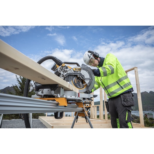 A construction worker wearing safety gear operates a DEWALT sliding compound miter saw (model DCS782) to cut a long wooden board at an outdoor building site with a scenic backdrop.