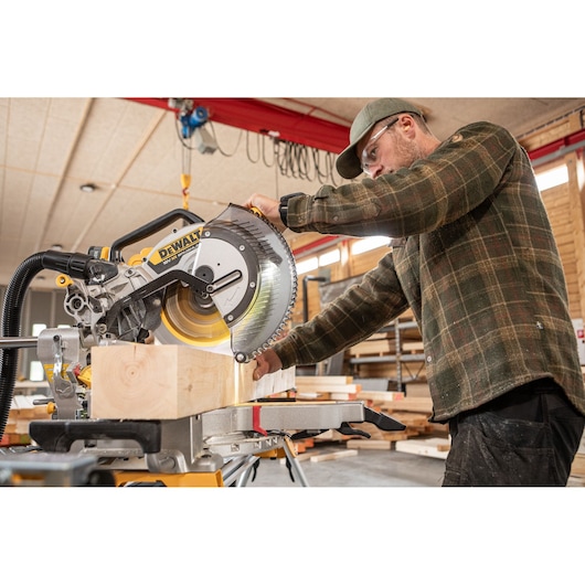 A person using a DEWALT DCS782N miter saw to cut a piece of wood in a workshop. The person's face is blurred for privacy.